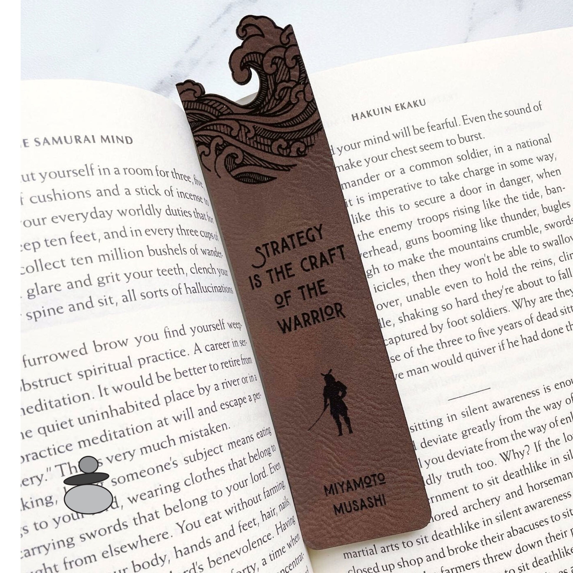 Brown Leatherette bookmark with cutout wave design on top. Below quote "strategy is the craft of the warrior" by Miyamoto Musashi with a samurai icon between quote and name.